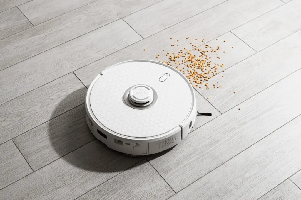 Белый робот-пылесос на светлом деревянном полу, убирающий разсыпанные крупы